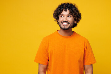 Wall Mural - Young smiling shocked surprised happy Indian man he wearing orange t-shirt casual clothes look aside on empty free area mock up isolated on plain yellow background studio portrait. Lifestyle concept.