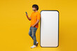 © ViDi Studio - Young smiling happy Indian man wear orange t-shirt casual clothes stand near big huge blank screen mobile cell phone with area using smartphone isolated on plain yellow background. Lifestyle concept.