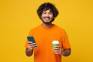 Wall Mural - Young smiling fun Indian man wear orange t-shirt casual clothes hold takeaway delivery craft paper cup coffee to go use mobile cell phone isolated on plain yellow background studio. Lifestyle concept.