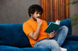 © ViDi Studio - Side view young Indian man wear orange casual clothes sit on blue sofa couch read book be in reverie mood stay at home hotel flat rest relax spend free spare time in living room indoor Lounge concept