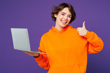 Young smiling happy Caucasian IT woman she wear orange hoody casual clothes hold use work on laptop pc computer showing thumb up isolated on plain purple background studio portrait. Lifestyle concept.
