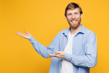 Wall Mural - Young smiling happy Caucasian man he wear blue shirt white t-shirt casual clothes point hands arms aside indicate on area mockup isolated on plain yellow background studio portrait. Lifestyle concept.