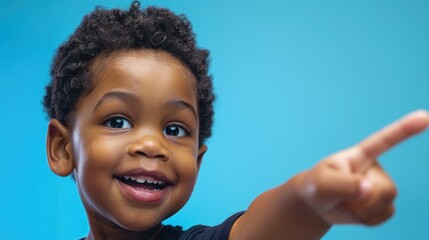 Close-up of a joyful young boy smiling and pointing to the side in a studio setting with a blue background. Happy child in casual attire