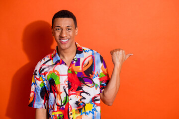 Wall Mural - Young man with cheerful expression pointing to side against bright orange background, wearing a trendy, colorful shirt, summer style