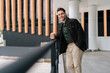 © dikushin - Vertical portrait of handsome bearded man holding takeaway coffee cup leaning on railing while standing by modern building, smiling looking at camera. Happy male drinking enjoying coffee outdoor