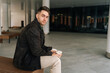 © dikushin - Portrait of stylish handsome young man sitting on bench in contemporary city environment, holding two coffee cup and looking at camera with serious expression. Concept of urban lifestyle.