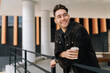 © dikushin - Handsome positive young man relaxing during coffee break, leaning on railing standing by modern office building. Happy bearded male in casual attire smiling looking away. Concept of urban lifestyle.