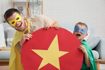 Canvas Print - Little boy and his father in superhero costumes with paper shield at home