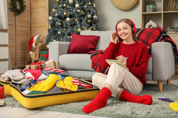 Wall Mural - Female traveler in headphones doing notes while packing suitcase at home on Christmas eve