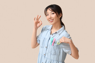 Wall Mural - Happy young Asian woman with clothespins showing OK gesture on beige background