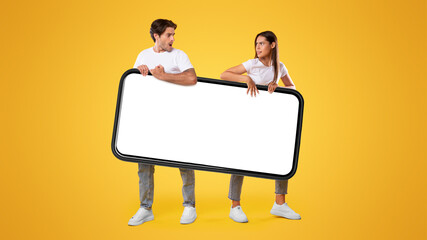 Poster - It's Mine. Couple holding big smartphone with white empty screen on yellow orange studio background fighting for gadget pulling device in different directions. Mock up display template, banner