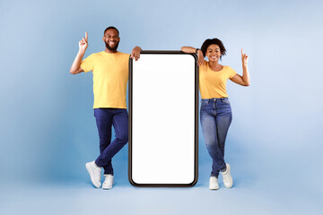 Poster - Cheerful African American couple standing next to big smartphone with blank white screen, pointing upwards, recommending new mobile application on blue studio background