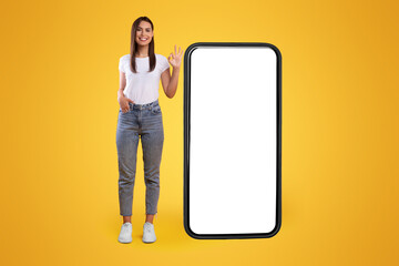 Aufkleber - Cool Mobile Offer. Happy casual lady standing near big huge cellphone with empty white screen, showing ok, recommending great new app or website for cell phone, orange wall, mock up, full body length