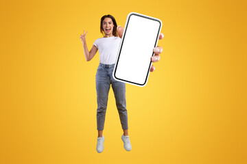 Wall Mural - Cool Mobile Offer Ad. Joyful young casual lady holding big empty cell phone screen jumping up in the air on orange studio background, showing peace sign gesture, free copy space, vertical