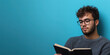 © dewaai - A young man wearing glasses, reading a book with a thoughtful expression, sitting casually with a blue background with copy space