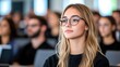 © trustmastertx - Young woman with glasses attentively listens in a lecture hall filled with other students.