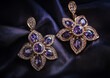 © Duka Mer - A pair of golden earrings with intricate flower-shaped designs, adorned with purple gemstones and diamonds, rest gracefully on dark satin fabric, exuding elegance and sophistication.