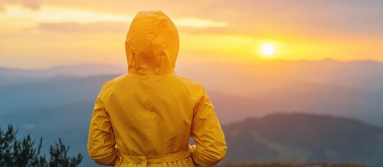 Wall Mural - Woman in yellow raincoat observing sunset over mountains with space for text and emotional reflection on nature's beauty.