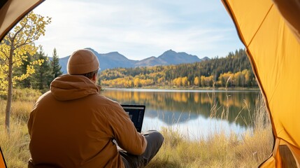 Photo from the back of Man in tent overlooking lake working on laptop, copy space. Concept: Digital Nomad, Remote Work, work and life balance, freelancing