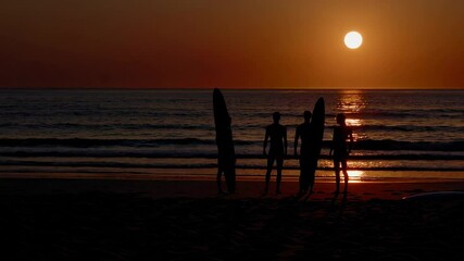 Poster - Silhouetted surfers standing together on sandy shoreline, holding surfboards while watching vibrant sunset sinking below ocean horizon in dramatic slow motion sequence