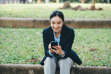 Poster - Professional Asian businesswoman smiling, checking smartphone, resting on low park wall during workday break, wearing tailored suit with confident demeanor