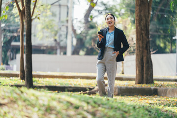 Poster - Professional businesswoman walking through urban park, scrolling smartphone while experiencing sunny day, representing career success and modern work-life balance