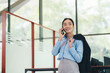© PaeGAG - Professional Asian businesswoman in blue shirt, gray skirt, holding jacket, speaking on phone and smiling confidently while standing in contemporary corporate environment