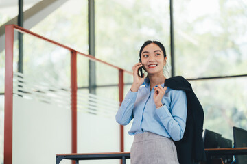 Poster - Professional Asian businesswoman in blue shirt, gray skirt, holding jacket, speaking on phone and smiling confidently while standing in contemporary corporate environment