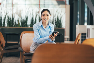 Poster - Professional Asian businesswoman sitting in sleek office lobby, texting on smartphone, wearing light blue shirt with gray skirt