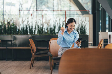 Poster - Asian businesswoman sitting on chair is using smartphone and celebrating success in modern office with happiness and excitement, business and technology concept