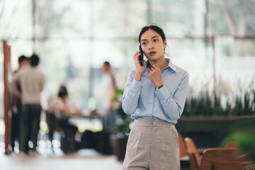 Poster - Professional Asian businesswoman talking on smartphone while walking through contemporary office, colleagues blurred behind, conveying busy corporate environment