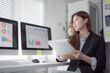 © Tj - Young businesswoman holding a document and analyzing financial charts and graphs displayed on computer screens, working with concentration and focus in a modern office environment
