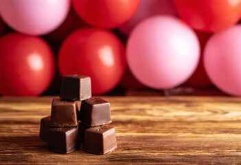 Wall Mural - An inviting photo featuring a collection of artisanal heart-shaped chocolates stacked on a rustic wooden table. In the background, a cascade of heart-shaped balloons in shades of pink and red creates 