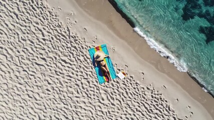 Poster - Top view of young woman in bikini and straw hat relaxing sunbathing on colorful towel on white sand beach with turquoise water gently lapping the shore in slow motion