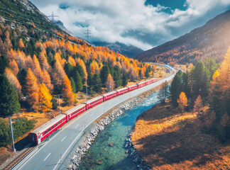 Wall Mural - aerial view of modern red train, river in alpine mountains, road, orange forest, sky with clouds at 