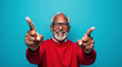 © OKA - Happy elderly Black man in glasses and red sweater pointing up against a blue backdrop. He has a bright smile, joyful facial expression, and raised index fingers, symbolizing enthusiasm and excitement