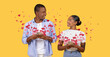 © Prostock-studio - A loving couple smiles at each other as they exchange messages on their smartphones. Surrounded by heart graphics, they celebrate Valentines Day with affection and connection.