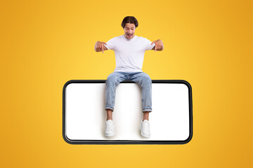 Wall Mural - Mobile App Advertisement. Handsome Excited Man Sitting On White Empty Smartphone Screen And Pointing Fingers Down Isolated Over Orange Studio Background. Check This Out, Cell Phone Display Mock Up