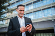© Jose Calsina - Confident mature businessman in formal suit standing outdoors using a cellphone to browse online. Business male focused on using smartphone for communication, checking emails, or managing work tasks