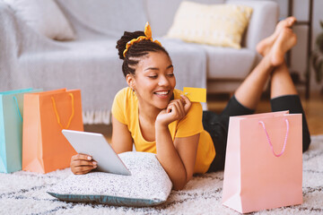 Poster - Buying Gifts And Presents Concept. Happy Black Lady Using Digital Tablet, Holding Credit Card, Ordering Goods In Internet Store, Lying On The Floor Carpet With Pillow, Looking At Shopping Bags