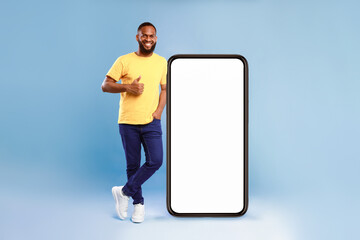 Poster - Cool mobile application. Joyful African American guy in gesturing thumb up near giant smartphone over blue studio background, mockup for website or new app on blank screen