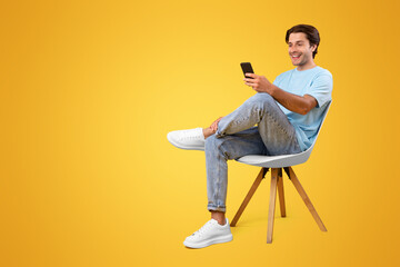 Poster - Portrait of excited smiling handsome millennial guy sitting on chair, using mobile application on modern smartphone, chatting with friends or watching funny video, orange background, vertical