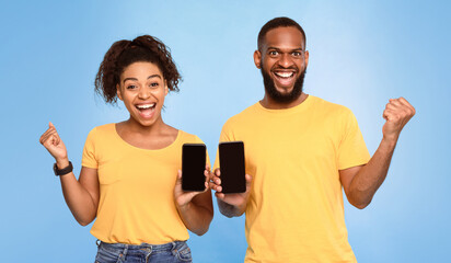 Poster - Amazing mobile app. Excited African American couple showing smartphones with mockup, gesturing YES, advertising new website over blue studio background. Big win, huge sale, great luck concept