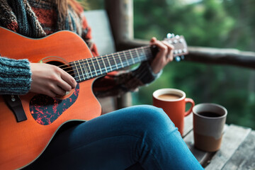 A woman in a cozy sweater strums an acoustic guitar on a porch, surrounded by nature and two coffee mugs, creating a serene and relaxing mood.