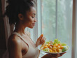 © Muhammad - A beautiful fitness woman in beige sports top eats healthy food near window, holding salad and meal with hands close up shot from the side, smiling happy looking at plate full of vegetables