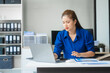 © Phushutter - Asian businesswoman sits at her ergonomic desk, working on a laptop and mobile phone. Her posture ensures minimal strain, addressing neck pain, back pain, and preventing repetitive strain injuries.