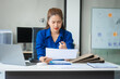 © Phushutter - Asian businesswoman sits at her ergonomic desk, working on a laptop and mobile phone. Her posture ensures minimal strain, addressing neck pain, back pain, and preventing repetitive strain injuries.