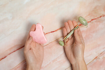 Wall Mural - Woman with face roller and gua sha tool on pink marble background, top view