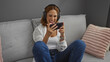 © Krakenimages.com - Young woman playing on smartphone wearing headphones while sitting on couch in cozy living room