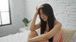 © Krakenimages.com - Young, chinese woman sitting on bed in bedroom, looking worried with hand on forehead, implying stress in a calm, indoor, home environment.